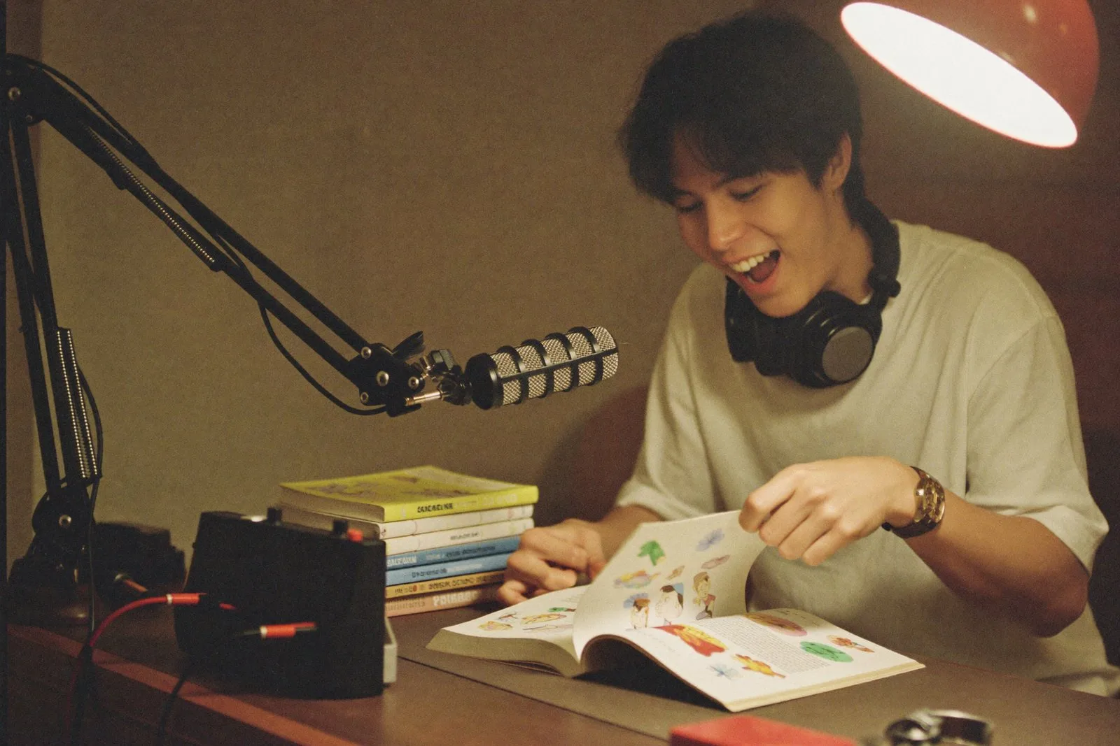 A young man with headphones around his neck in a small home studio with a microphone and a stack of picture books