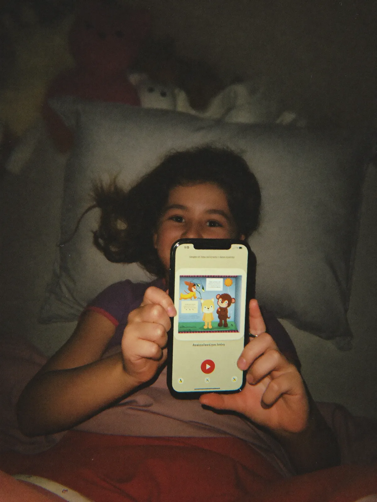 A five-year-old in bed with the lamp low, holding a phone in both hands and listening to a story
